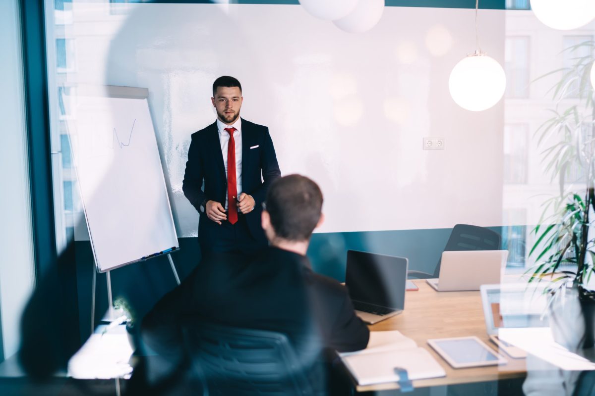 Through glass of concentrated male worker in formal wear standing near whiteboard and explaining business plan to colleagues in office